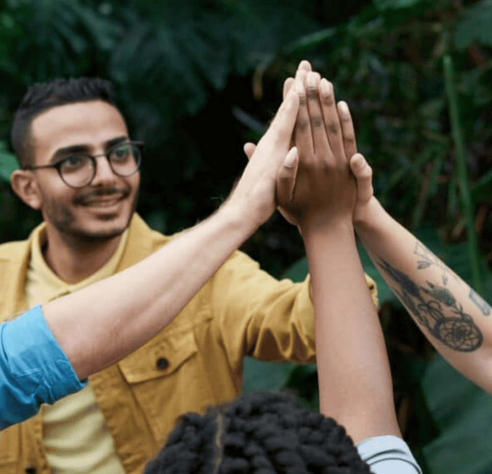 A group of four people standing outdoors in a lush green environment, giving each other a high-five. One person is wearing glasses and a yellow jacket, another has tattoos on the arm, and the others are partially visible.