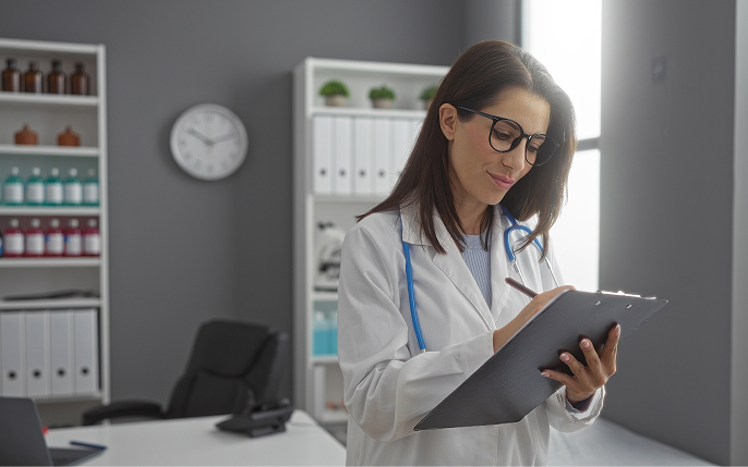 A female medical professional wearing glasses, a white lab coat, and a blue stethoscope looks down intently while writing on a clipboard. The background shows a clean, modern medical office with organized shelves and a desk.