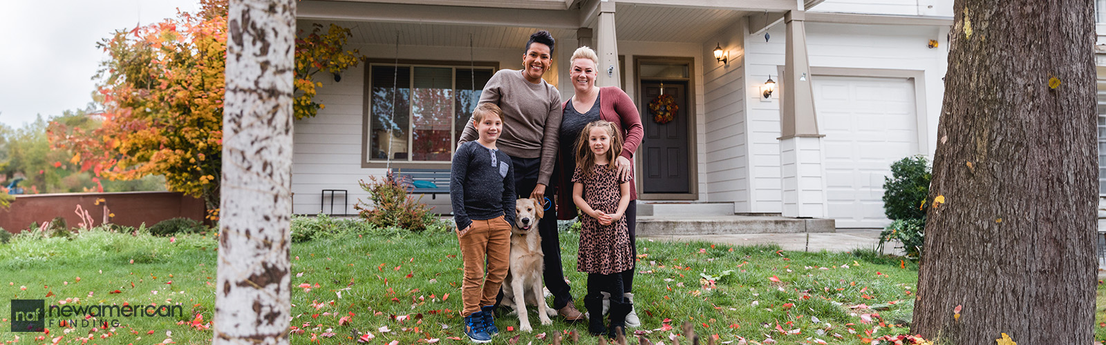 An interracial lesbian couple stands smiling with their two kids in front of their new home