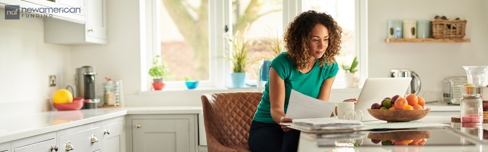 A woman with brown skin and curly hair stands at a kitchen island going over paperwork in front of her laptop