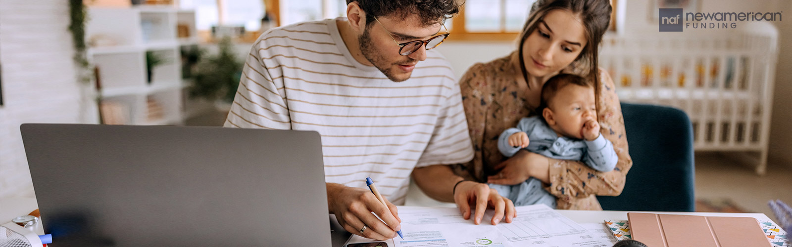 A white man and woman sit at a table looking at financial forms while the woman holds a baby