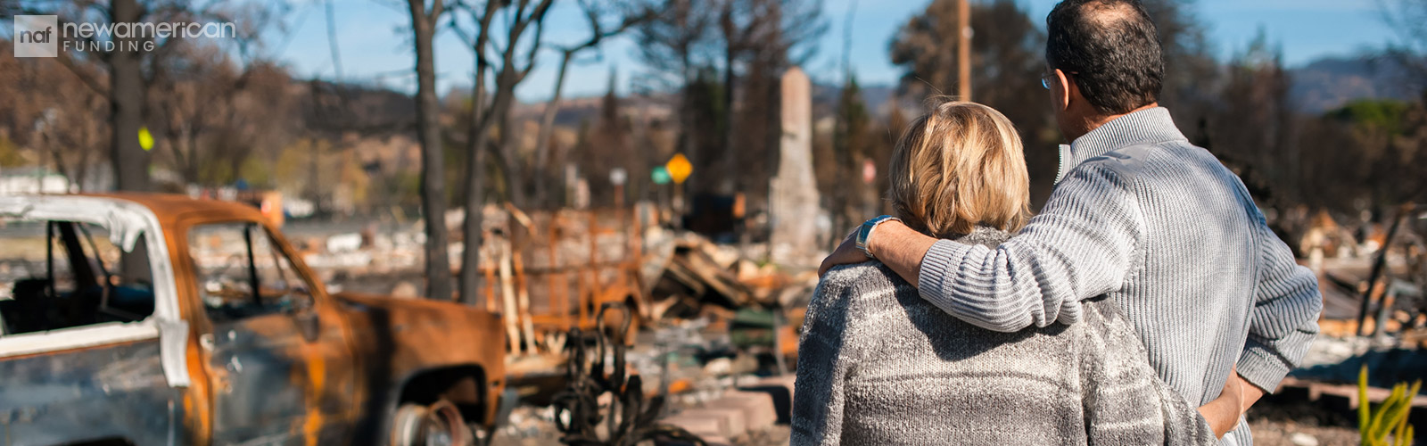 A man and woman stand with their arms around each other looking at the remains of their home
