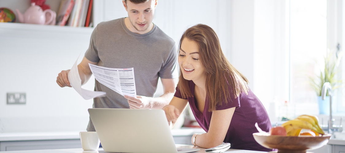 A white couple leaning over a kitchen counter holding up paperwork and looking at an open laptop and smiling.