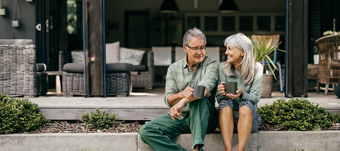 A senior white woman and man sit laughing together on the stairs to their home