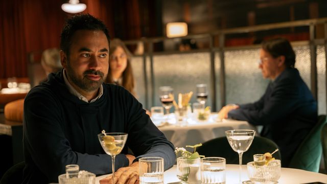 Kal Penn as Jay Jonah Atterbury sits at a restaurant table with a martini, watching others in a dimly lit dining room.