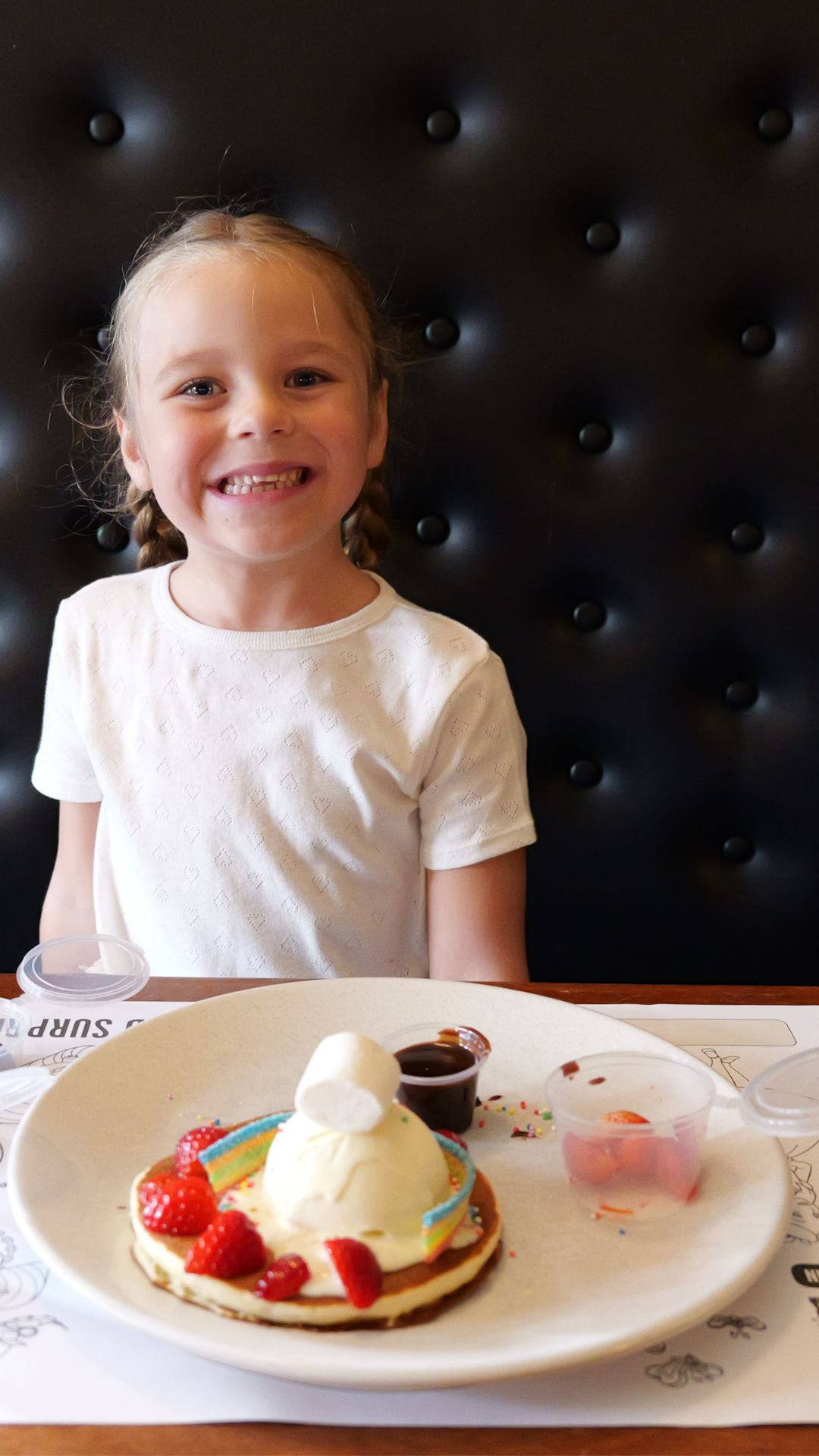 A child sitting in a restaurant booth decorating a pancake topped with ice cream, strawberries, marshmallows and colourful toppings, with small containers of sprinkles and sauce on the table.