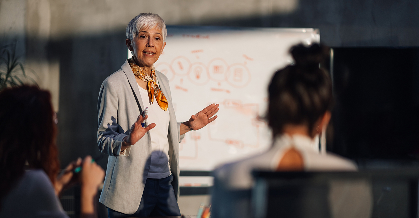 A confident older woman with short grey hair, wearing a blazer and a patterned scarf, stands in front of a whiteboard and gestures while giving a presentation to a small group in an office with strong sunlight.