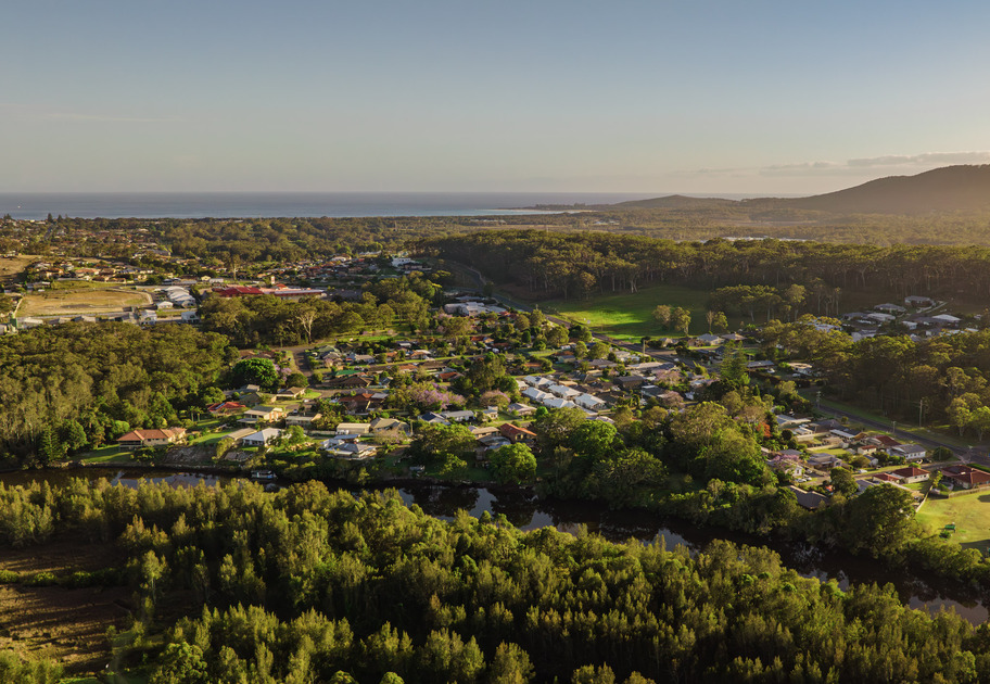 The sun sets over a rural landscape, casting a warm golden light on green hills, scattered trees, and a small cluster of buildings under a clear sky
