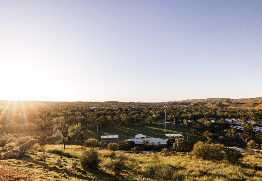 A scenic view of a grassy hill overlooking a sports field and buildings, surrounded by trees and low mountains, with the sun setting on the horizon and casting a warm glow over the landscape.