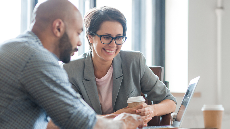 A man and a woman are looking at a laptop screen together in a bright office. The woman is wearing a gray blazer and glasses, holding a coffee cup, and smiling at the screen. The man's back is to the camera. This suggests a collaborative or mentoring session.