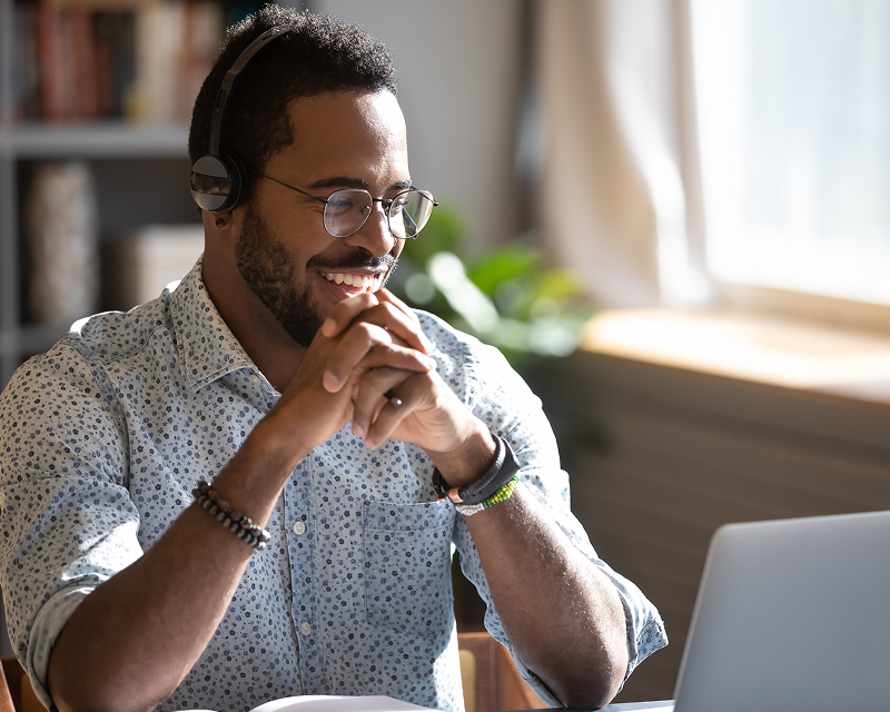 A smiling man wearing glasses and over-ear headphones is sitting at a desk with a laptop open in front of him. He is wearing a light blue patterned shirt and has bracelets on his wrist, suggesting he is on a video call or engaged in online learning.