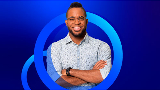 A smiling man with glasses, a patterned button-down shirt, and a stylish haircut stands with his arms crossed. The background is a bright blue with concentric blue circles.