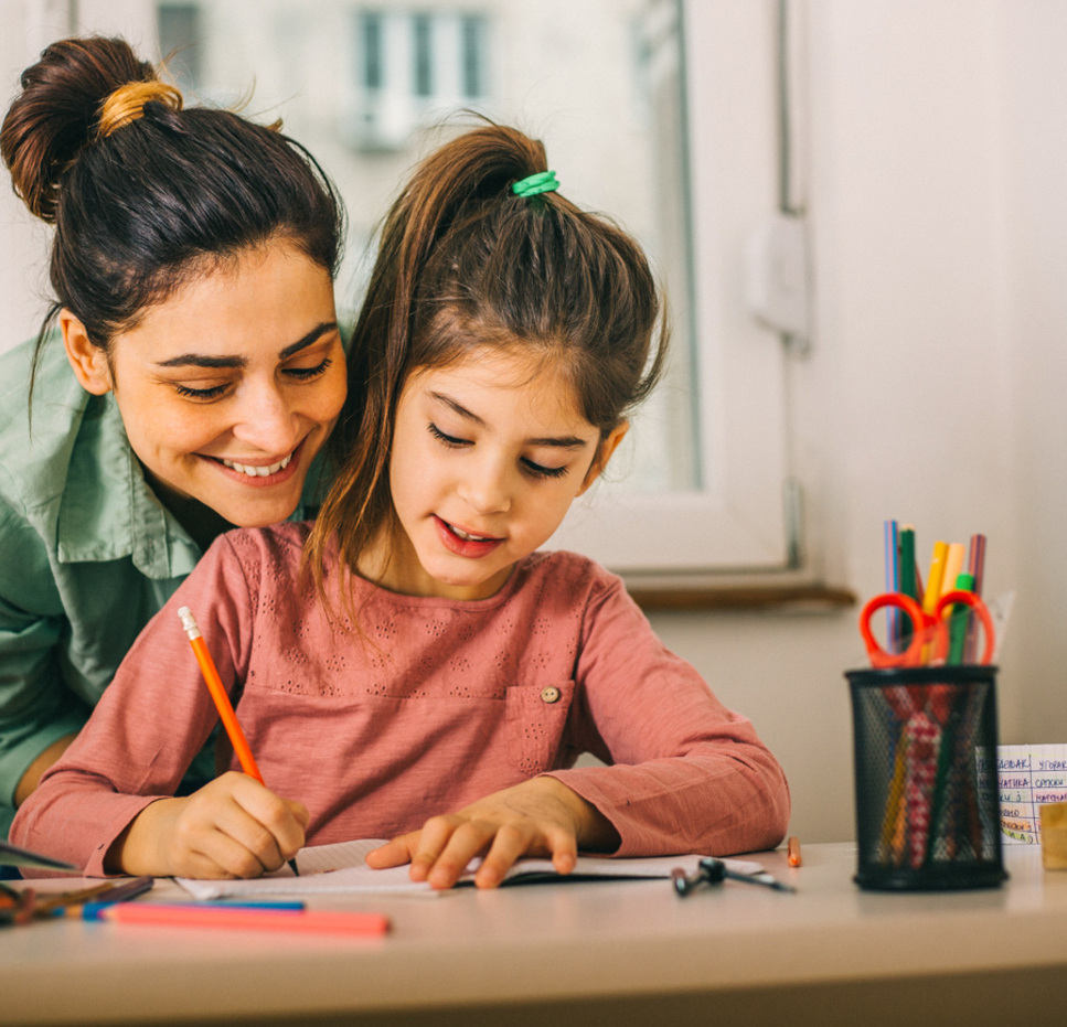 A woman smiles and leans over to help a young girl with her homework at a desk, surrounded by pencils, colored pens, and a Connect Four game. The girl is writing in a notebook while the woman guides her.