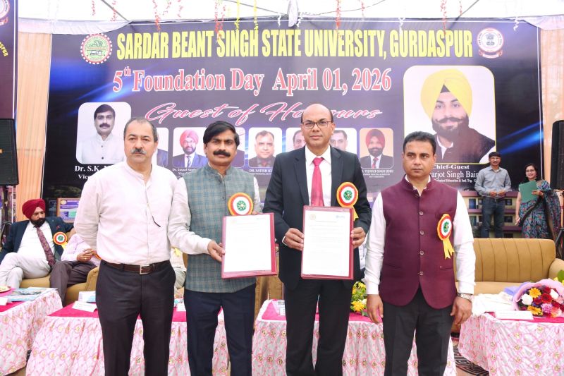 Four men stand in the foreground of a stage, holding up documents in red folders. They are positioned in front of a large banner that reads 'SARDAR BEANT SINGH STATE UNIVERSITY, GURDASPUR' and commemorates the '5th Foundation Day April 01, 2026.'