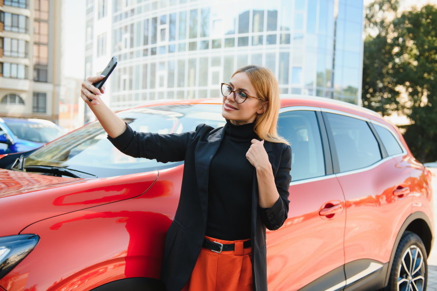 Mlada ženska ponosno pozira za sebek ob vzorno bočno parkiranem vozilu.