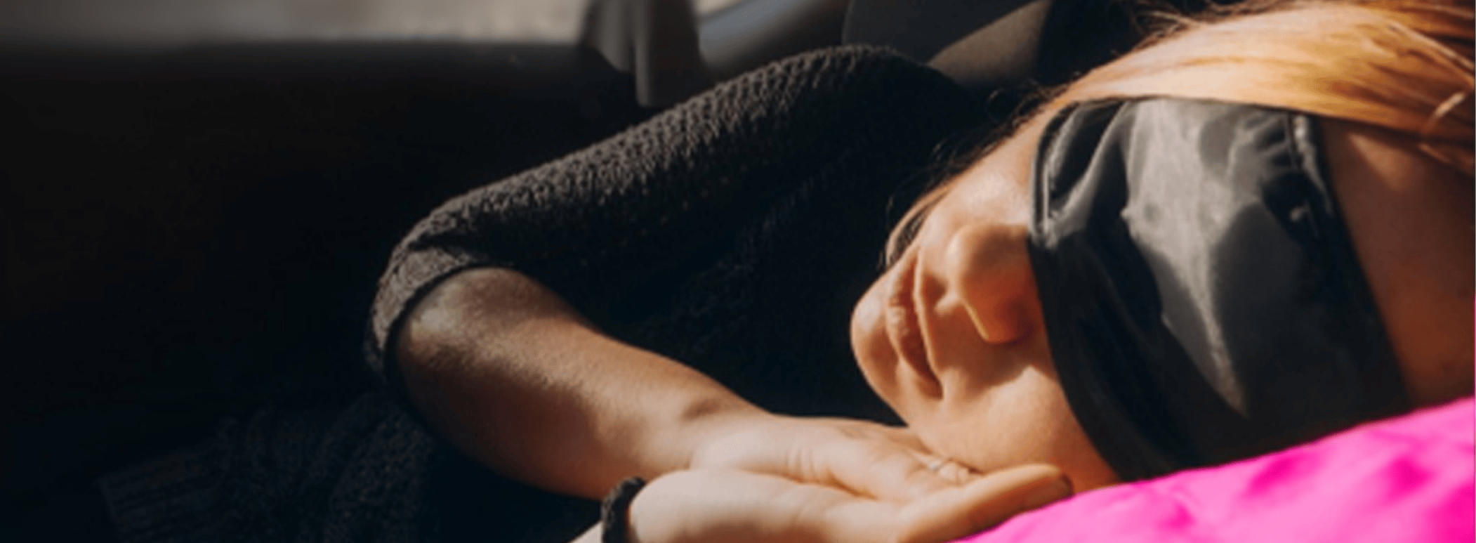 Person resting in their car while wearing a sleep mask, lying on their side with a bright pink blanket.