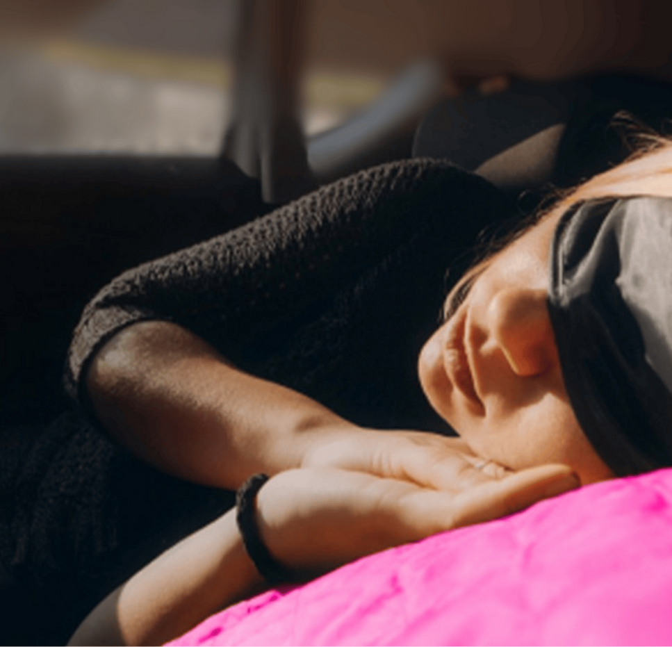 Person resting in their car while wearing a sleep mask, lying on their side with a bright pink blanket.