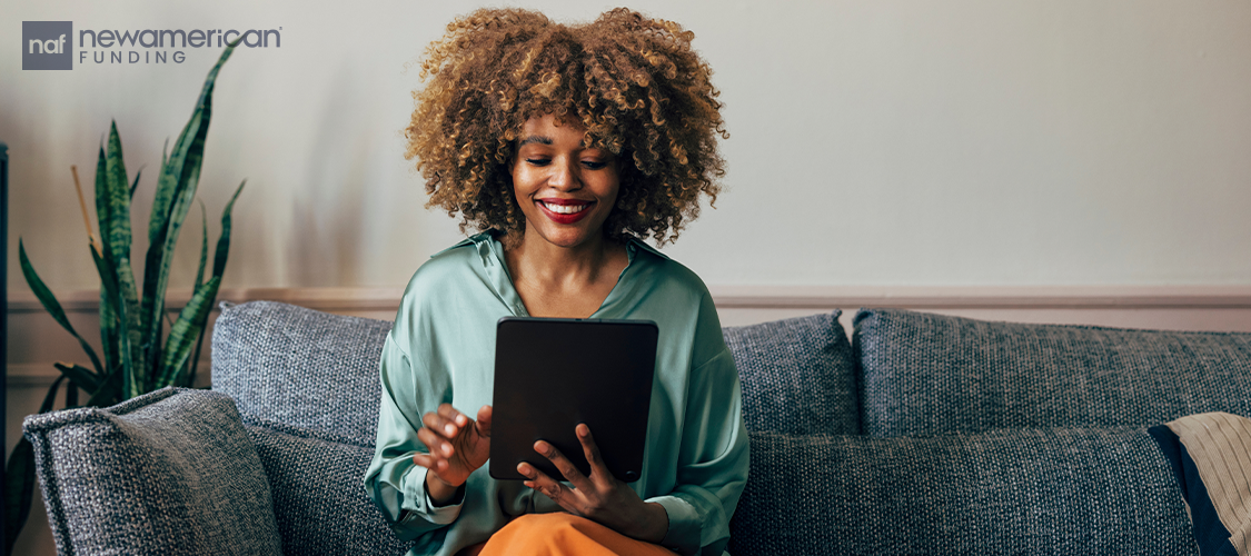 A woman sitting on a couch smiling as she holds a tablet.