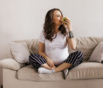 Mlada nosečnica sedi na kavču po turško in se pripravlja, da bo zagrizla v jabolko.