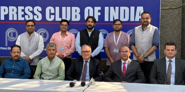 A group of ten men pose for a formal photograph at the Press Club of India. Five men are seated behind a long table covered in a white cloth with two microphones placed in the center, while five others stand behind them. They are positioned in front of a large blue backdrop prominently featuring the 'Press Club of India' name and logo repeated across the top. The group is dressed in a mix of formal suits and traditional Indian Nehru jackets.