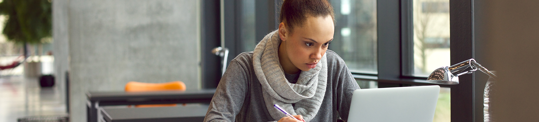 A woman wearing a gray sweater and a chunky gray scarf sits at a desk, looking intently at a silver laptop and writing on a piece of paper. The setting is a modern office space with large windows and concrete walls.