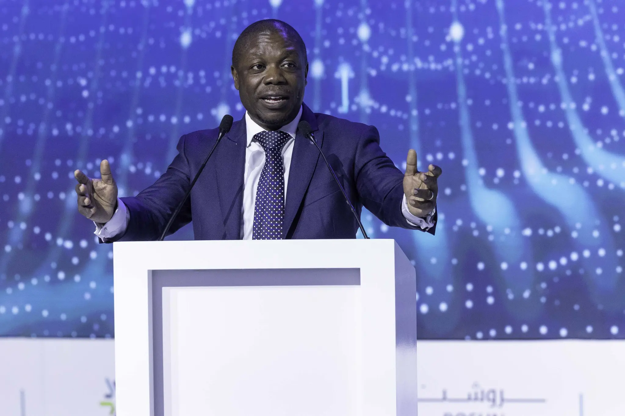 Man in a suit speaking at a CIPS conference podium with blue digital background.
