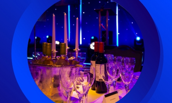 Event table setup with wine bottles, glasses, and candles against a stage backdrop with blue lighting