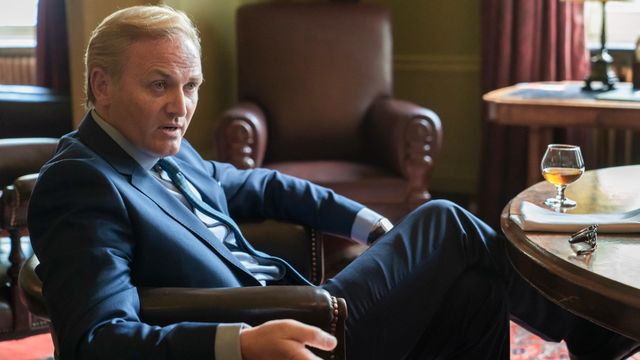 Roger Barclay as Otto Mostyn in a leather chair, gesturing as he speaks to someone out of frame
