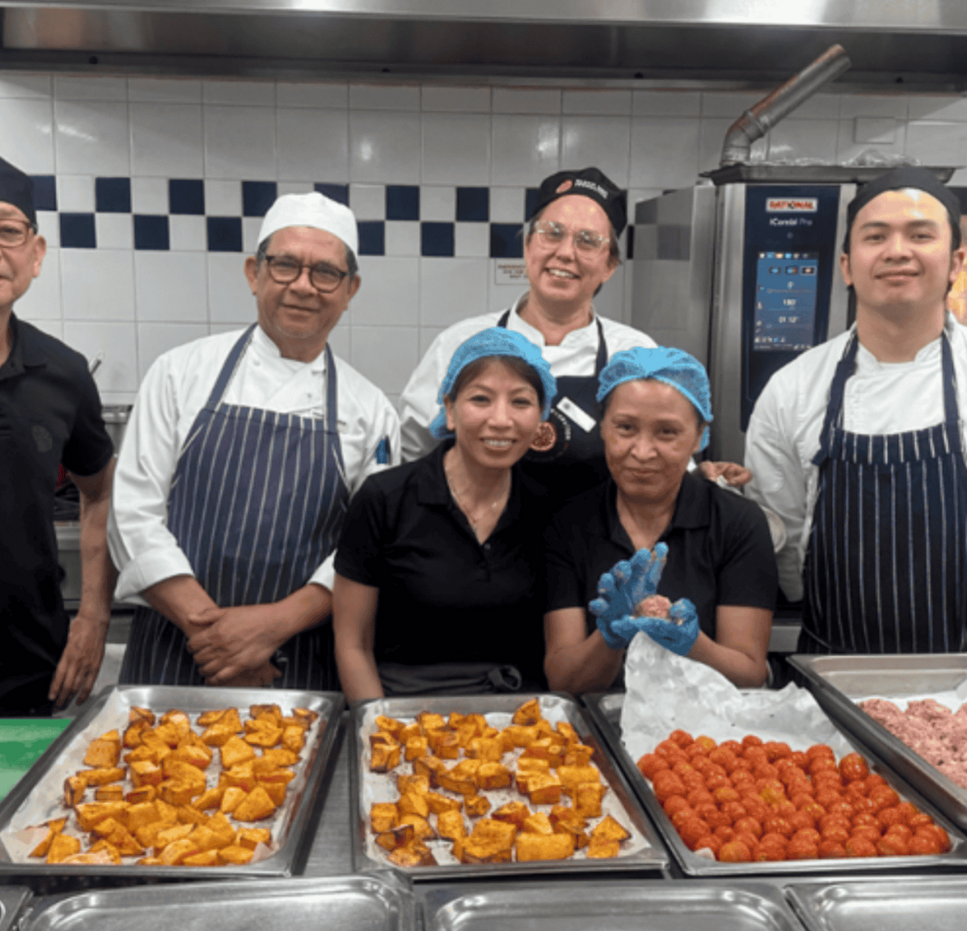 Mission Australia aged care chefs and kitchen staff smiling together in the kitchen, standing behind trays of freshly prepared ingredients including pumpkin, tomatoes and meatballs.