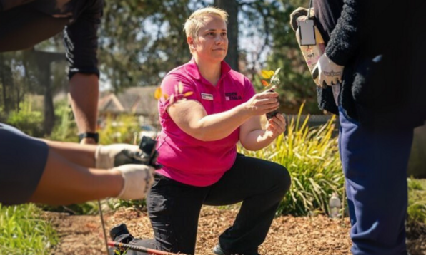 A person in a pink shirt kneels on the ground while holding a small plant. The person is surrounded by others who are also engaged in gardening activities. The background shows greenery and a house.
