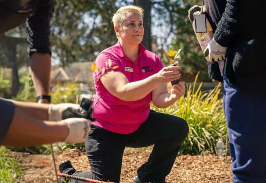 A person in a pink shirt kneels on the ground while holding a small plant. The person is surrounded by others who are also engaged in gardening activities. The background shows greenery and a house.