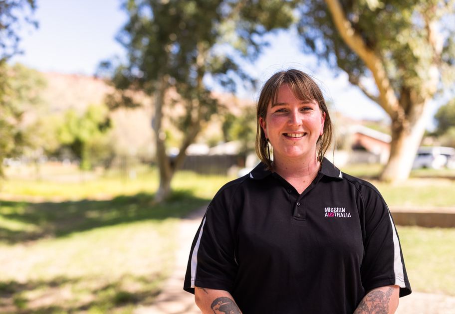 A person with shoulder-length hair and tattoos smiles outdoors, wearing a black Mission Australia polo shirt. Sunlight filters through trees in the background, creating a bright, natural setting.



