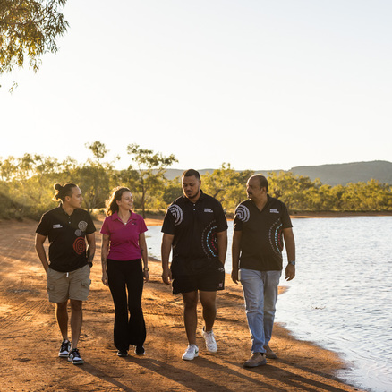 Four people walk together along a lakeside dirt path in the countryside, talking and smiling, with trees and hills in the background under a clear sky.