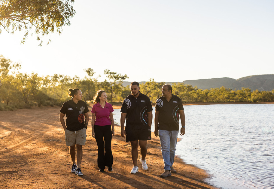 Four people walk together along a lakeside dirt path in the countryside, talking and smiling, with trees and hills in the background under a clear sky.