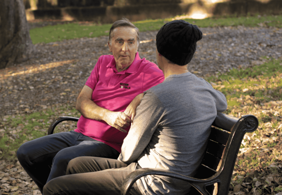 Two people sit on a park bench having a conversation; one wears a pink shirt, and the other wears a gray shirt and black beanie.