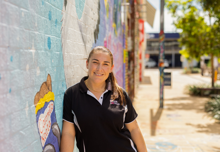 A Mission Australia case worker with long brown hair in a ponytail, wearing a black and white polo shirt, stands smiling against a colorful mural on a sunny sidewalk. Trees and buildings are visible in the background.