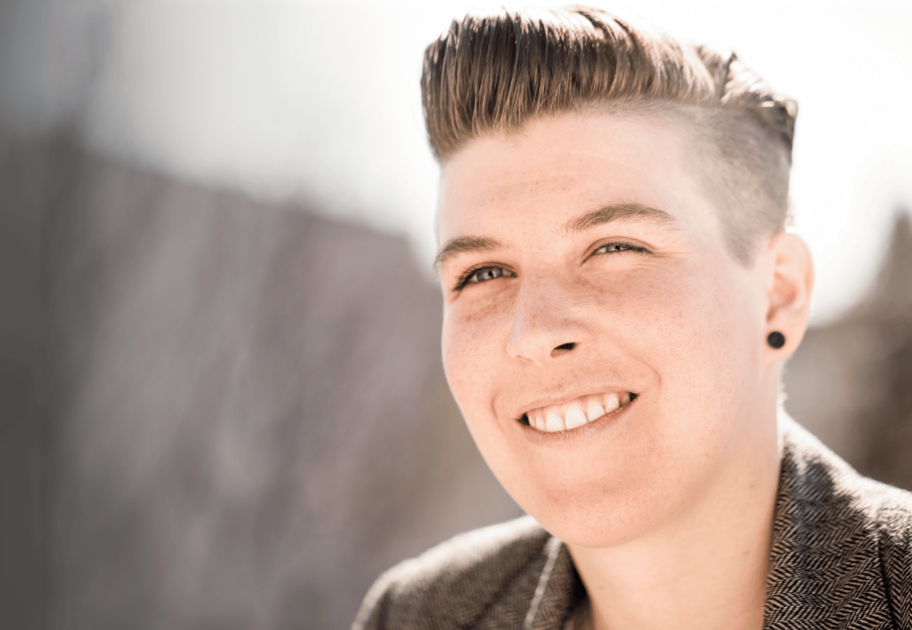 Smiling person with short styled hair and earrings, wearing a patterned jacket, standing outdoors in bright sunlight.