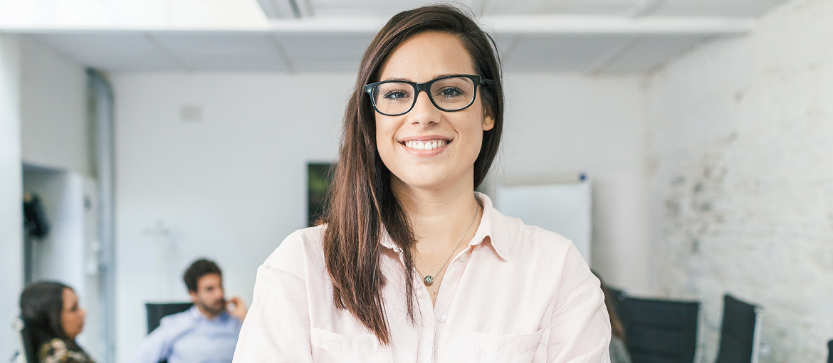 A young woman with glasses and long dark hair, wearing a pink shirt, smiles directly at the camera in a bright office meeting room. Other people are seated in the blurred background.