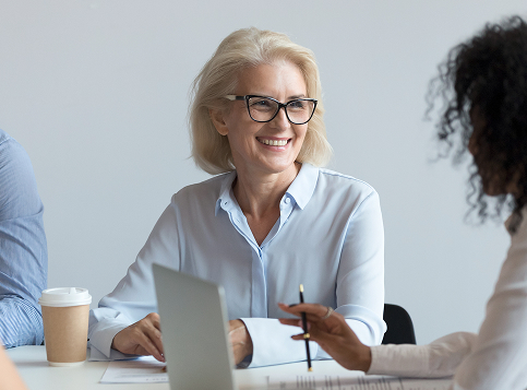 A smiling, mature woman with blonde hair and glasses sits at a table across from a colleague during a meeting. A laptop, documents, and a coffee cup are on the table.