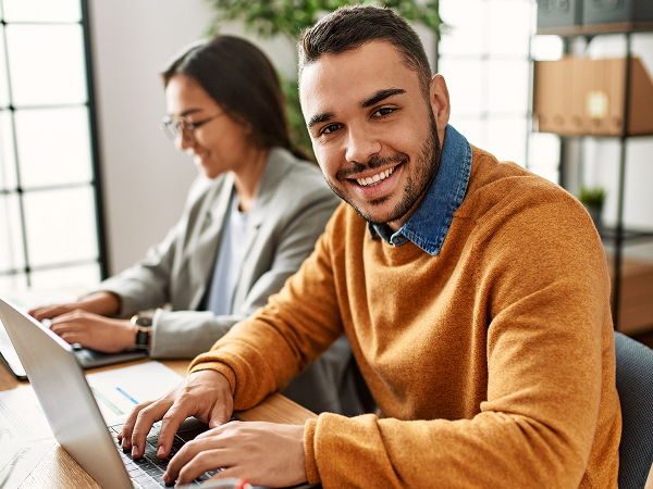 A young man in a mustard-colored sweater smiles at the camera while typing on a laptop in an office. A woman is blurred and working on her laptop behind him.