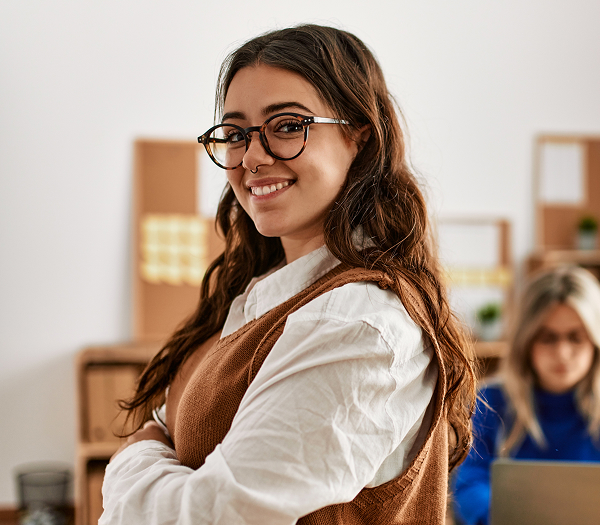 A young woman with long wavy hair, glasses, and a brown vest smiles confidently at the camera, standing in a brightly lit office. A person works in the blurred background.