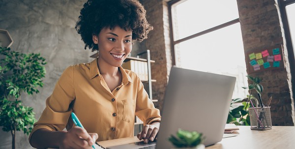 A young woman with an afro hairstyle, wearing a mustard-yellow shirt, smiles while looking at a laptop and writing in a notebook at an office desk.
