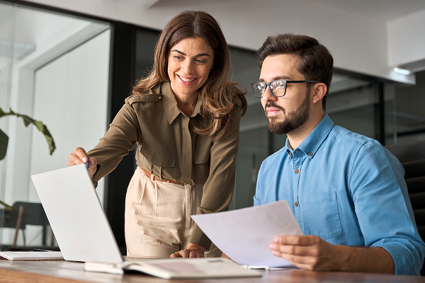 A woman in a khaki shirt points to a laptop screen while standing next to a man in a blue shirt who is seated at a desk holding papers. They are collaborating in an office.