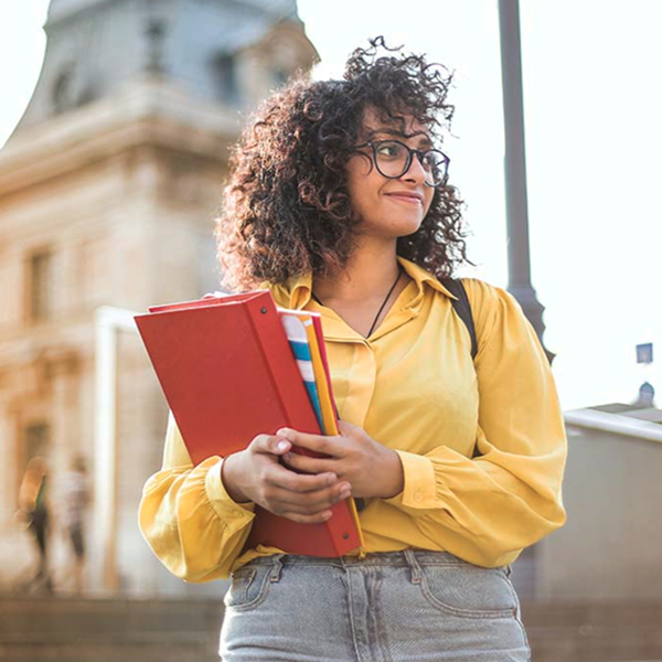 A young woman with curly hair and glasses, wearing a bright yellow shirt, stands outdoors holding red binders and looking to the side. A stone building is visible in the background.