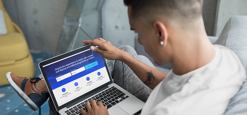 Over-the-shoulder shot of a person sitting on a couch, using a laptop that displays a website with the search phrase 'Search for somewhere to study, take exams and find an accredited programme provider'