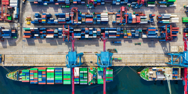 An aerial top-down view of a busy shipping port featuring two large container ships docked at a pier, with giant blue and red cranes loading multicolored cargo containers.