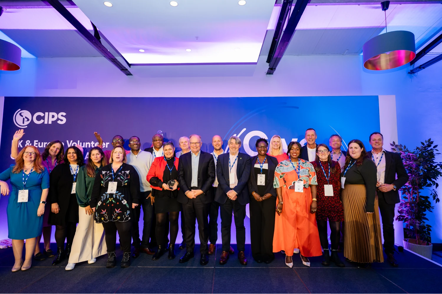 A large group of people, dressed in business and formal attire, posing for a group photo on a stage. They are standing in front of a blue CIPS UK & Europe Volunteer Conference and Awards 2025 banner. Several individuals are holding awards.