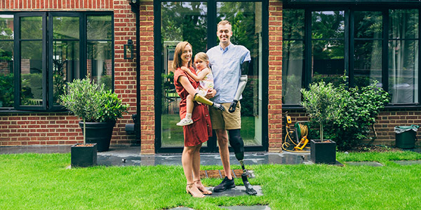 A disabled couple stands smiling with their child in front of their new home