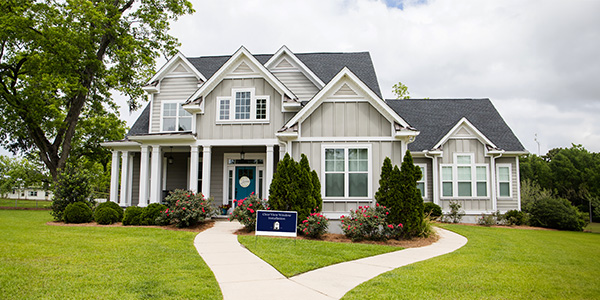 A nice two story house with grey siding sits on a well-manicured lawn with a large, beautiful tree beside it