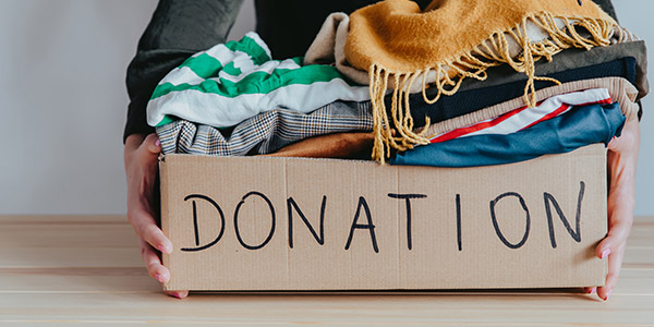 A pair of hands holds a cardboard box filled with clothes with the word donation written in sharpie on the side